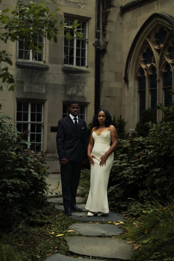 A couple in formal attire on a stone path framed by greenery and gothic windows