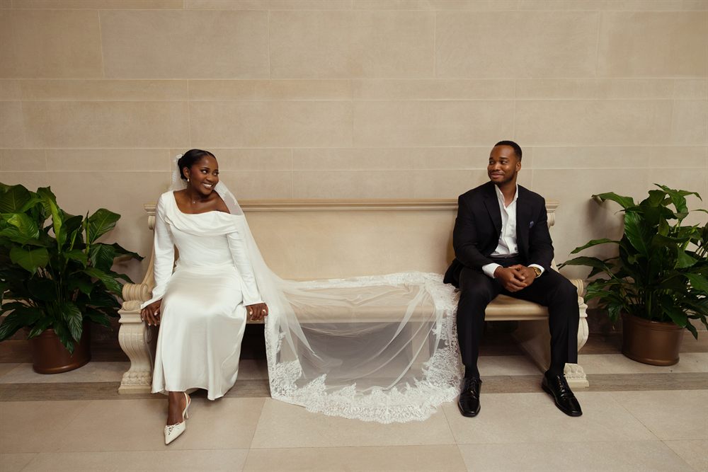 A bride and groom smiling at each other from either end of a stone bench at the National Gallery of Art, her lace veil spread between them
